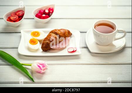 Sandwich croissant frais avec jambon, œufs, tomates et radis et une tasse de thé sur table blanche. Petit déjeuner sain et romantique servi avec amour et tulipe rose Banque D'Images