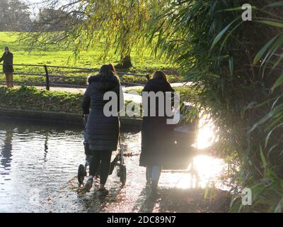 Deux jeunes mamans en manteau d'hiver ont silhoueté dans la lumière du soleil d'automne, poussant leurs prams sur le chemin inondé au bord du lac à Abington Park. Banque D'Images