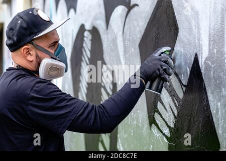 Graffiti artiste en action, dessin sur le mur avec aérosol peinture dans une boîte, portant masque de protection / respirateur avec filtres. Banque D'Images