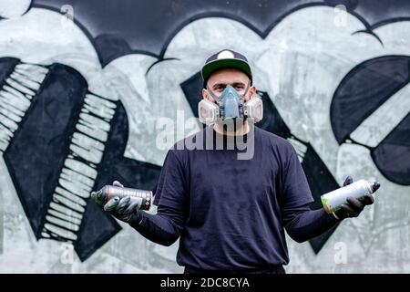 Graffiti artiste posant devant son dessin sur le mur, avec deux peintures aérosol dans une boîte, portant un masque facial / respirateur de protection. Banque D'Images