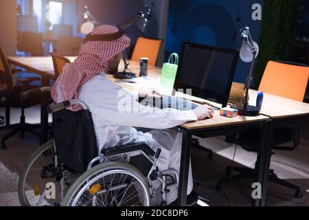 Homme d'affaires arabe handicapé dans la tradition suit travaillant en fonction pendant la pandémie de coronavirus. Homme d'affaires handicapé dans le fauteuil roulant travaille en ligne i Banque D'Images