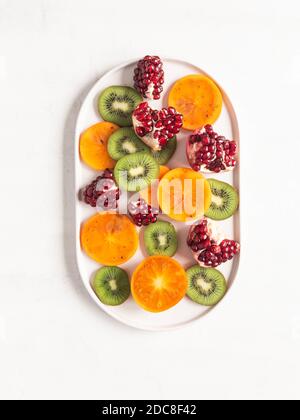 Fruits naturels sains de saison multicolores - persimmon, kiwi, tranches de grenade sur plaque ovale blanche. Vue du dessus. Espace de copie de la mise à plat Banque D'Images