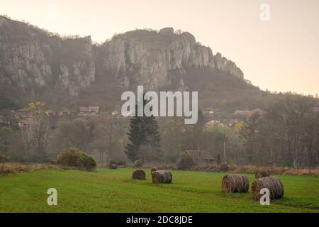 Village pittoresque de Vlasi dans le canyon de la rivière Jerma, au pied d'une falaise rocheuse massive et de balles de foin en premier plan sur un champ vert Banque D'Images