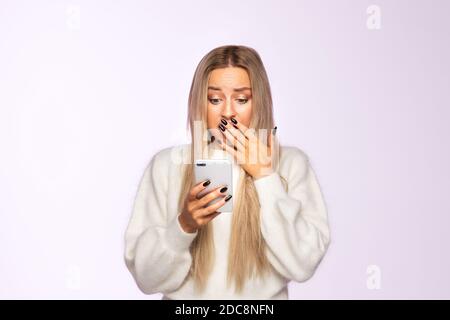 Surprise jeune femme couvrant la bouche avec la main et regardant son téléphone, reçoit des sms, posant sur fond violet doux. Émotions humaines, conc. Réaction Banque D'Images