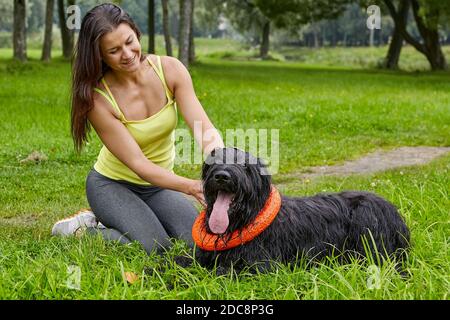 Black briard est couché sur l'herbe avec sa femme propriétaire souriante dans le parc public. La femme et le chien à poil long se reposent pendant la marche. Banque D'Images