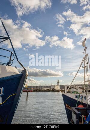 Un phare et un quai sont aperçus entre les arcs de deux chalutiers qui font face l'un à l'autre. Un ciel bleu est au-dessus avec des nuages. Banque D'Images