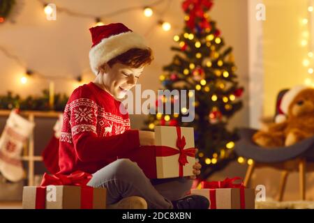 Positif garçon dans des vêtements de fête rouges assis et déballer des boîtes Avec des cadeaux de Noël Banque D'Images