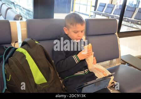 Un garçon s'assoit à une gare ou à une aroport en attendant un train ou un avion dans sa veste, en mangeant un sandwich ou en jouant un comprimé. Petit sac à dos garçon Banque D'Images