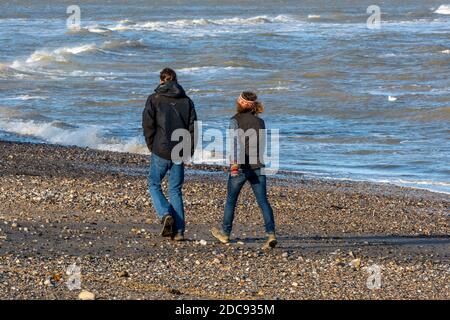 un homme et une femme marchant le long d'une plage pierreuse lors d'une journée venteuse portant des manteaux imperméables. Banque D'Images