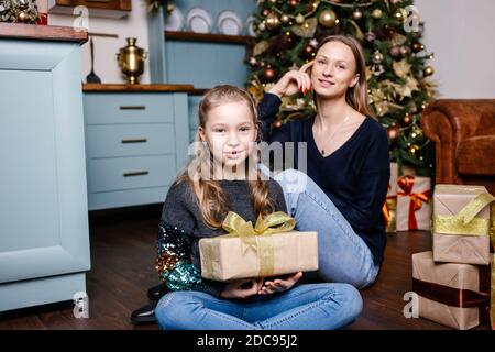 La mère va faire la surprise pour la fille donne le présent, près de l'arbre de Noël à la maison. Une fille souriante heureuse reçoit un cadeau de sa mère. Banque D'Images