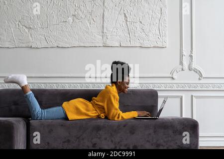 Souriante femme afro-américaine biraciale avec une coiffure afro-américaine dans un gilet jaune allongé sur un canapé, au repos, regardant la webcam de la caméra et parlant sur la vidéo cha Banque D'Images