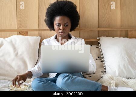 Femme afro-américaine concentrée en chemise blanche se détendant après le travail, mangeant du pop-corn tout en regardant un film d'horreur ou de thriller sur un ordinateur portable, allongé au lit. Paresseux r Banque D'Images