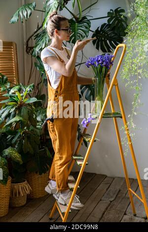 Femme souriante fleuriste dans des combinaisons orange debout sur escabeau, prend une photo de bouquet d'iris pourpre dans vase en verre sur smartphone. Amour pour la fleur Banque D'Images