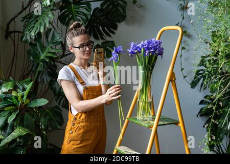 Femme souriante fleuriste dans des combinaisons d'orange prend une photo de bouquet d'iris pourpre dans un vase en verre sur un smartphone, debout sur un escabeau. Amour pour la fleur Banque D'Images