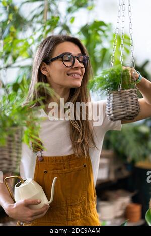 Femme jardinière en combinaison orange arrosage maison en pot en serre entourée de plantes et de pots, en utilisant blanc arrosoir métal. Maison gardeni Banque D'Images