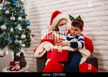 Un garçon de 5 ans excités avec une prop sur sa tête assis sur les genoux du Père Noël avec un masque facial et ouvrant un cadeau de Noël à côté de l'arbre de Noël. Hiver Banque D'Images