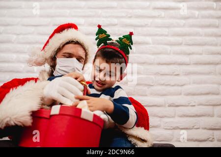 Un garçon de 5 ans souriant et enthousiaste avec un soutien sur lui Tête assise sur les genoux du Père Noël avec masque et observation Un cadeau de Noël rouge à côté d'un arbre de Noël Banque D'Images