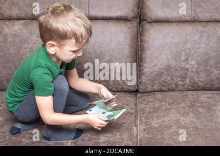 Enfant caucasien concentré dans des vêtements décontractés tient cent euros dans ses mains et compte de l'argent sur le canapé dans la chambre, vue latérale, espace de copie. C Banque D'Images