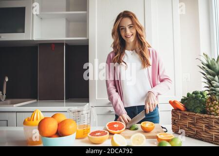 Freckles Ginger Woman sourit à l'appareil photo tout en coupant des fruits et préparer du jus Banque D'Images