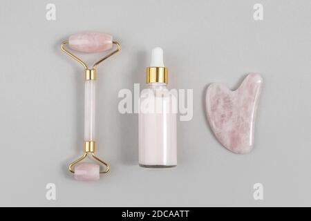 Rouleau facial en quartz rose cristal, outil de massage Gua sha et collagène anti-âge, sérum en bouteille de verre sur fond gris. Massage du visage naturel Banque D'Images