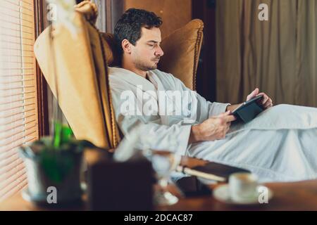 Un homme dans un peignoir blanc travaillant sur sa tablette À la chambre d'hôtel pendant COVID-19 Banque D'Images