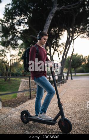 Jeune homme sur un kick scooter en jeans, à l'écoute de la musique dans la rue, attendant le bus. Banque D'Images