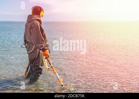 Un homme debout au niveau du genou dans l'eau à la recherche de métaux précieux avec un détecteur de métaux. Mer et ciel en arrière-plan. Lumière du côté droit. Son Banque D'Images