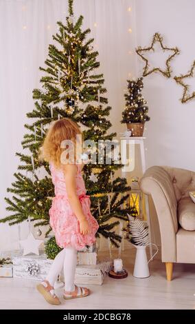 Fille de 6 ans méconnaissable en robe de fête rose, accrochée aux ornements de boules de Noël sur l'arbre de Noël. Salon blanc minimaliste. Banque D'Images