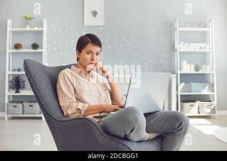 Femme sérieuse regardant l'appareil photo tout en travaillant sur un ordinateur portable assis dans un fauteuil confortable Banque D'Images