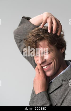 Jeune homme beau souriant positivement et avec assurance posant avec les mains sur son visage et sa tête, en regardant satisfait, amical et heureux contre le gris Banque D'Images