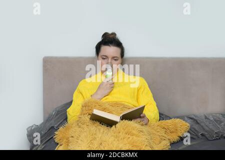 Femme caucasienne malade en chandail inhalation avec un nébuliseur à la maison sur le lit et lire le livre. Photo de style de vie. Banque D'Images