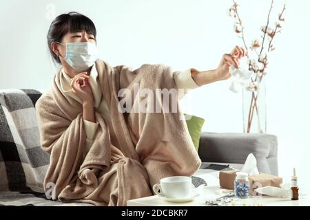 Femme enveloppée d'un tissu écossais, portant un masque facial essayant de se protéger des malades à proximité, semble déguisée, en colère, triste assis sur le canapé à la maison à l'intérieur. Soins de santé et médecine, prévention des maladies. Banque D'Images