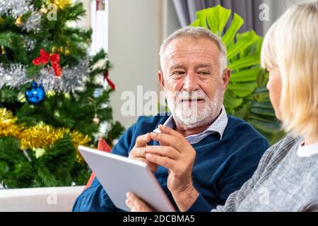 Couple senior utilisant une tablette à un appel vidéo pour accueillir leur famille pour le festival de Noël, assis sur un canapé avec décoration et arbre Banque D'Images