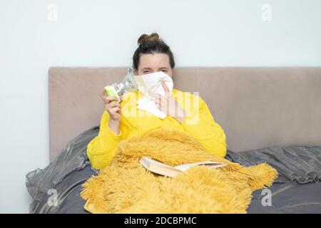 Une femme fatiguée et malade au lit dans un chandail et sous une couverture dans son livre de lecture à la maison, serviette en papier et nubélizer sur l'auto-isolation pendant Banque D'Images
