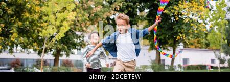 Enfant souriant tenant un cerf-volant coloré tout en courant près d'un ami asiatique dans le parc, bannière Banque D'Images