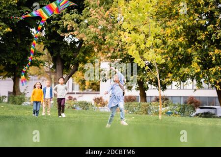 Fille jouant avec le cerf-volant tandis que des amis multiethniques marchent sur pelouse herbacée sur fond flou Banque D'Images