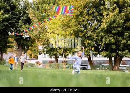 Enfant qui court avec un cerf-volant près de ses amis sur un arrière-plan flou sur la prairie dans le parc Banque D'Images