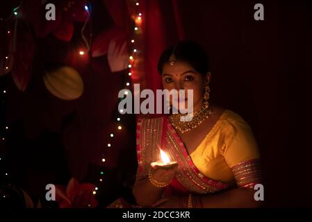 Une jeune et belle femme bengali indienne en robe traditionnelle indienne tient une diya/lampe Diwali dans sa main devant des lumières bokeh colorées. JE Banque D'Images