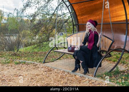 Une dame gaie est assise sur une balançoire de jardin, forgée dans un manteau bordeaux et le biret, avec de beaux yeux, à l'automne sur le fond d'un étang là Banque D'Images