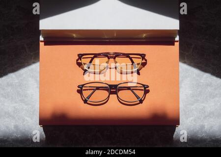 Deux verres d'ordonnance différents reposent sur un grand livre avec une couverture orange et une autre copie grise dans la lumière du soir sur un fond texturé gris. Banque D'Images
