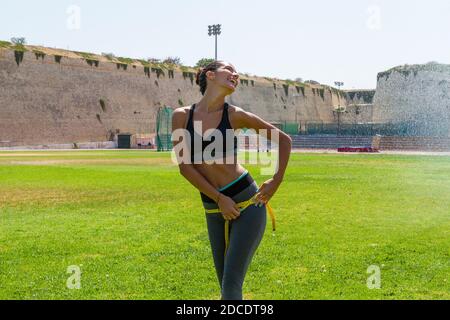 Femme athlète humide arrosée femme dans des vêtements de sport mesurant sa taille, ses hanches et sa poitrine après un entraînement dans un stade de terrain extérieur. Li sain Banque D'Images