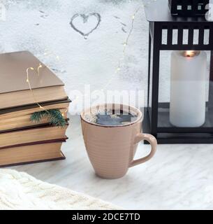 Pile de livres avec branche de sapin utilisé comme signet, grande tasse avec café noir frais, lanterne avec une bougie sur le seuil de fenêtre. Banque D'Images