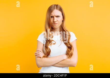 Portrait d'une jeune fille bouleversée, dans un T-shirt blanc, debout avec les mains pliées et le visage offensé, sur fond jaune Banque D'Images