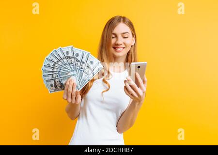 Portrait d'une femme excitée en t-shirt blanc montrant de l'argent factures et tenue de téléphone mobile isolé sur fond jaune Banque D'Images