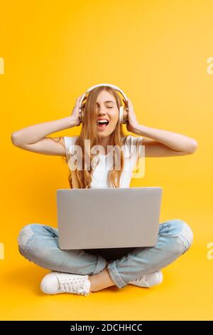 Une image d'une belle femme dans un T-shirt blanc écouter de la musique sur un casque à l'aide d'un ordinateur portable lorsque vous êtes assis sur fond jaune Banque D'Images