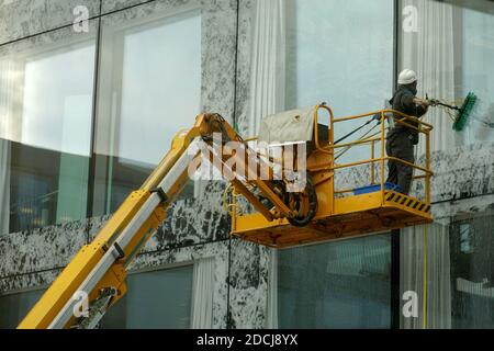 Homme lavant les fenêtres et la façade d'un bâtiment avec une vadrouille, il est debout sur une plate-forme de travail aérienne. Il porte une ceinture de sécurité et un casque. Banque D'Images