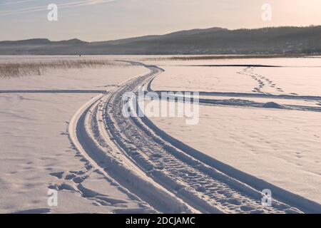 Une piste de motoneige mène au loin le long de la neige blanche du lac Big gelé au coucher du soleil. Banque D'Images