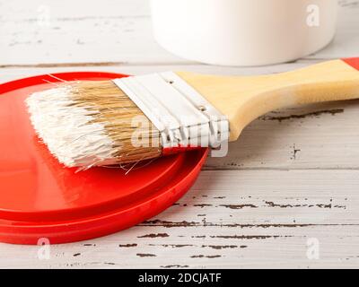 Nouvelle brosse à poils naturels avec peinture blanche sur un couvercle en plastique rouge sur une surface en bois rayée. Travaux de peinture de construction, réparation et redécoration. Banque D'Images
