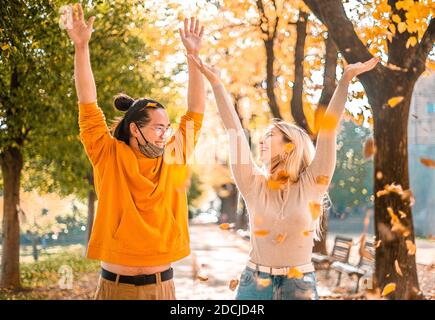 Couple souriant heureux avec masque de visage vers le bas, lancer automne automne feuilles rouges orange dans la forêt de parc à l'extérieur en temps de coronavirus - nouveaux cactus saisonniers normaux Banque D'Images