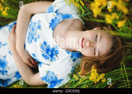 Belle mignonne souriante jeune femme caucasienne posant souriant sur l'herbe dans le champ d'été de fleurs dans une robe multicolore, vie saine Banque D'Images